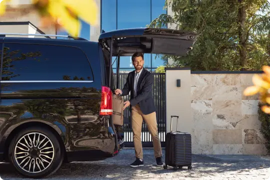 A man loads luggage into the back of a luxury black van outside a modern residence, highlighting relaxed, upscale travel moments.