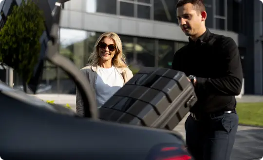 A chauffeur loads a suitcase into a luxury vehicle as a smiling woman stands nearby outside a modern building.
