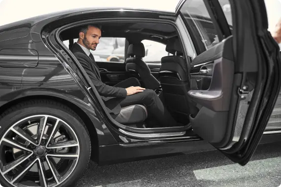 A man in formal attire seated in a luxury sedan with the door open, parked near a private jet terminal.