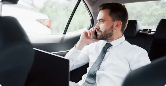 Businessman in a white shirt and tie taking a call while working on a laptop in the backseat of a luxury car.