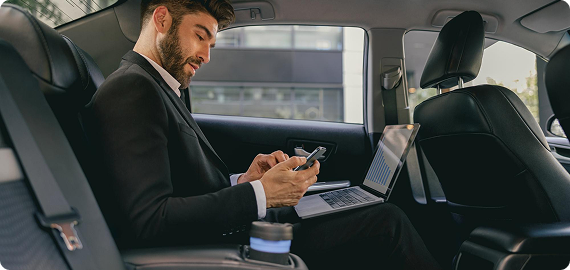 Businessman in a suit multitasking with a smartphone and laptop while traveling in the back of a luxury vehicle, coffee cup in foreground.