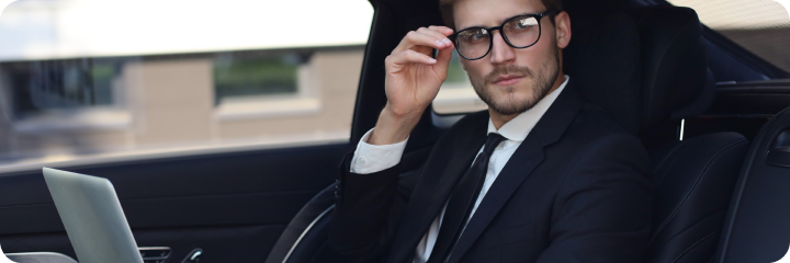 Young executive with glasses using a laptop in the backseat of a high-end black car, adjusting his eyewear and looking out the window.