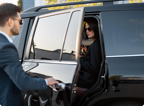 Female passenger in black attire and sunglasses stepping out of a luxury SUV as a chauffeur opens the door for her.