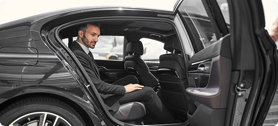 Man in a suit sitting in the back of a luxury sedan with the door open, preparing for departure.