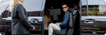 Businessman in sunglasses seated inside a luxury black SUV with sliding door open, looking out at a woman approaching the vehicle.