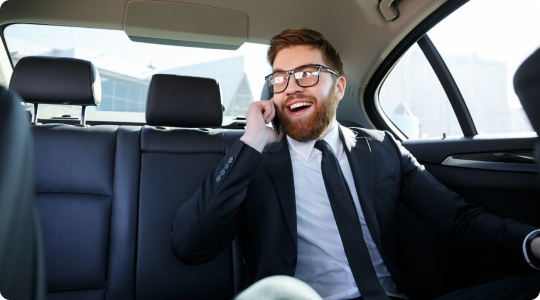 Young businessman smiling and talking on the phone while seated in the back of a premium sedan during daylight.