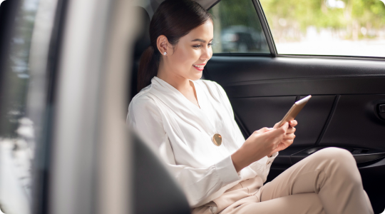 Young woman seated comfortably in the back of a vehicle, smiling while using a smartphone during daytime.