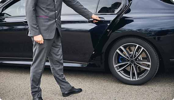 Man in a gray suit opening the back door of a high-end black sedan, positioned near the rear wheel on an asphalt road.