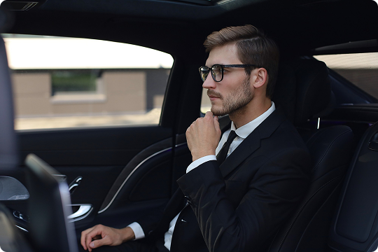 Young businessman with glasses seated in the back of a luxury car, resting his hand on his chin in a thoughtful pose.