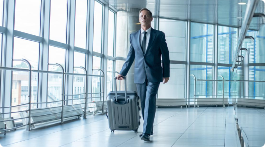 Businessman in a formal suit walking confidently through a modern airport terminal, pulling a wheeled suitcase.