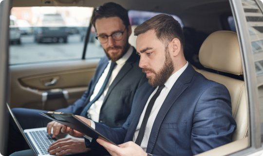 Two businessmen in suits reviewing documents on a clipboard and laptop while seated in the back of a premium car.