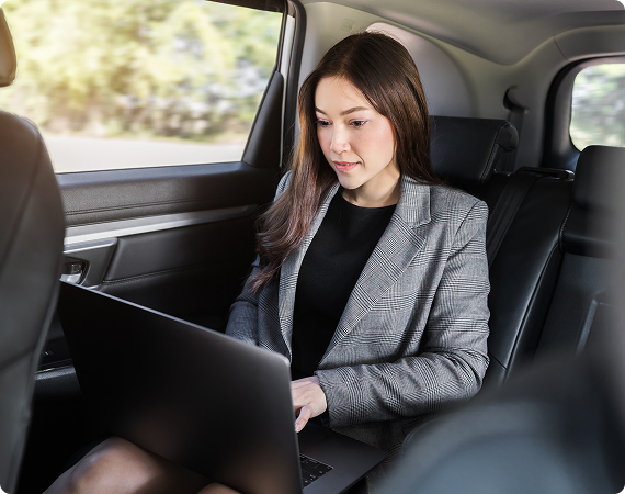 Businesswoman in a plaid blazer working on a laptop in the backseat of a car, focused and productive during transit.