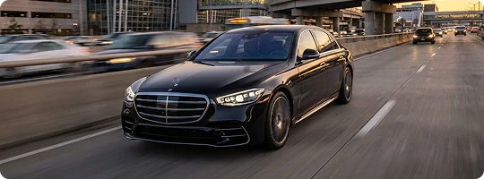 A black Mercedes-Benz S-Class in fast motion on a highway in sunset scene with other cars visible in blur motion in background having an overhead bridge and glass buildings.