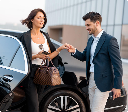 A stylish woman in a black blazer and a white top greeted by a man in a navy jacket and brown pants beside a luxury black car.