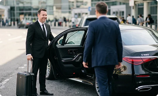 A smiling chauffeur in a suit holds open the door of a black luxury sedan while holding a suitcase handle. A male passenger viewed from behind approaches the vehicle at an airport terminal pickup area.