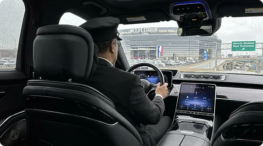A chauffeur in a uniform and cap drives a luxury car toward MetLife Stadium, visible through the windshield on a cloudy day.
