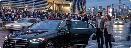 A couple stands by a black Mercedes Benz  with a chauffeur opening its rear door. The car is parked outside a stadium with its entrance filled with crowd and fast moving cars in blur motion as well as distant view of city buildings in evening.