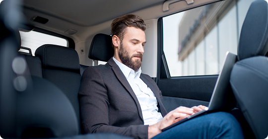 Bearded man in a dark blazer and white shirt working on a laptop in a spacious car backseat.
