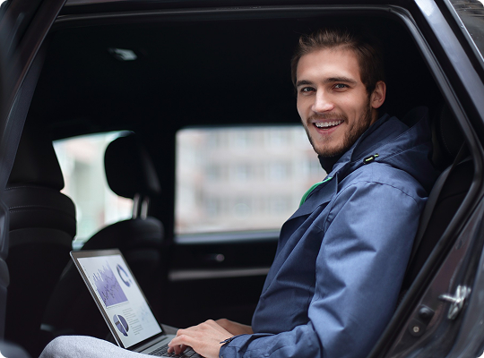 A smiling man in a blue jacket is reviewing graphs on a laptop while seated in the rear of a car.