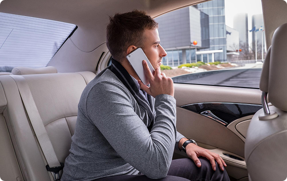 Man in casual attire making a phone call while seated in the backseat of a beige-interior sedan, with city buildings visible outside the window.