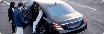The chauffeur assists the passenger exiting the black car at the airport parking area.