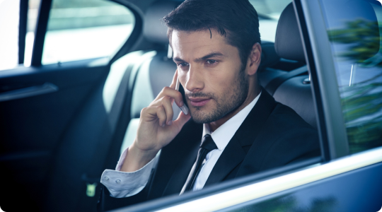 Serious-looking businessman in a suit talking on a smartphone while seated in the backseat of a luxury car, gazing out the window during transit.