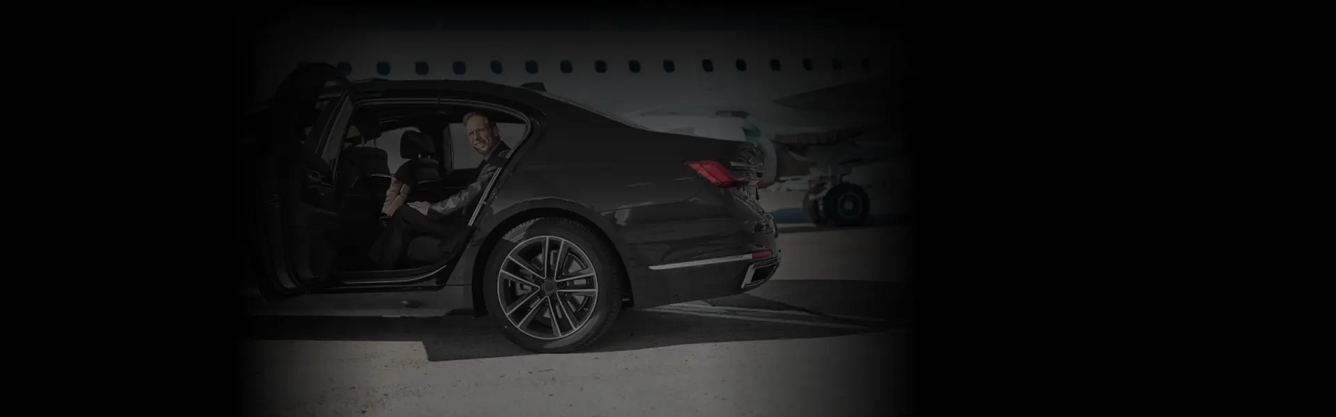 A man smiling inside a luxury sedan parked by a jet, showcasing a rear-angle view for LaGuardia Airport Transfers.
