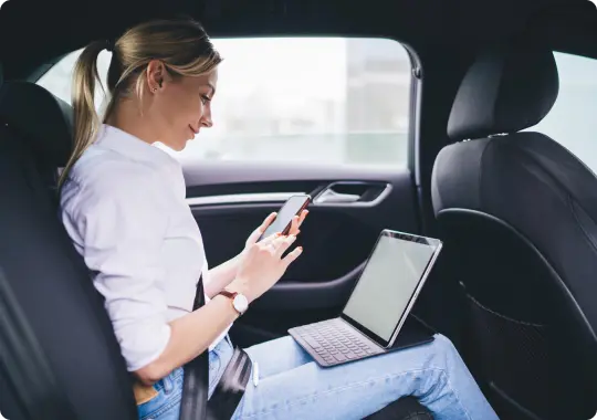 A young woman works comfortably from the backseat, using her phone and tablet mid-ride in a sleek executive vehicle.