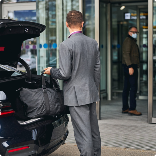 Man in a gray suit loading a large black duffel bag into the trunk of a luxury car outside an airport entrance, with a masked traveler visible in the background.