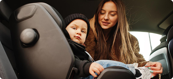 Mother buckling her toddler into a safety seat in the back of a premium car, with soft daylight streaming in.