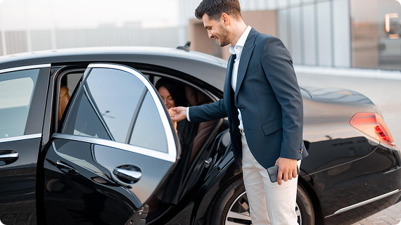 A chauffeur courteously opens a black car door for a smiling woman seated inside, framed in a cityscape background.
