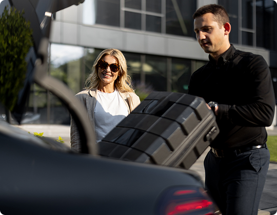 A chauffeur assisting with luggage while a smiling passenger looks on, framed from a low rear trunk angle for a polished travel vibe.